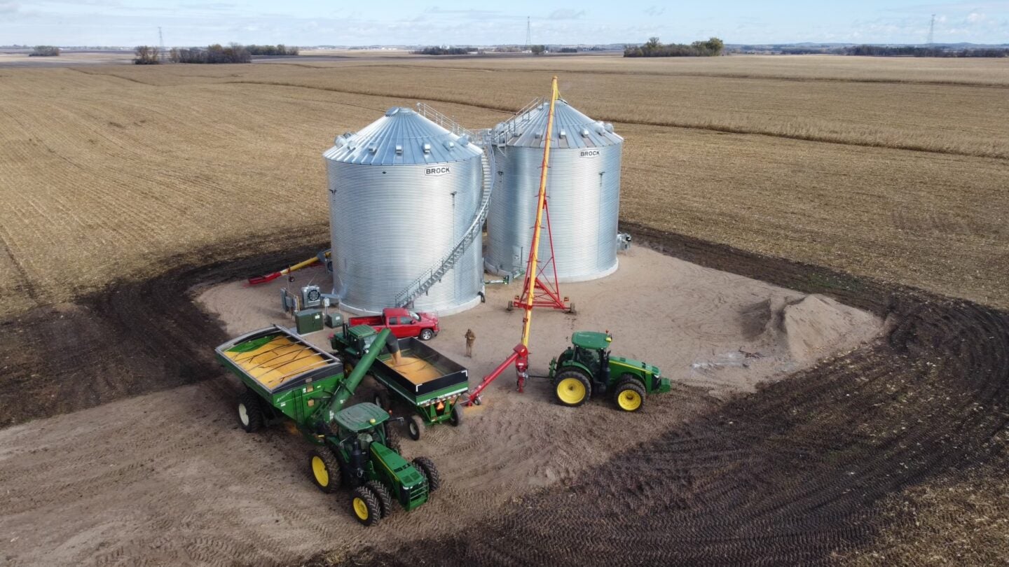 aerial view of grain silos on farmland with two tractors, one with a grain cart and the other with an auger. The surrounding land appears plowed