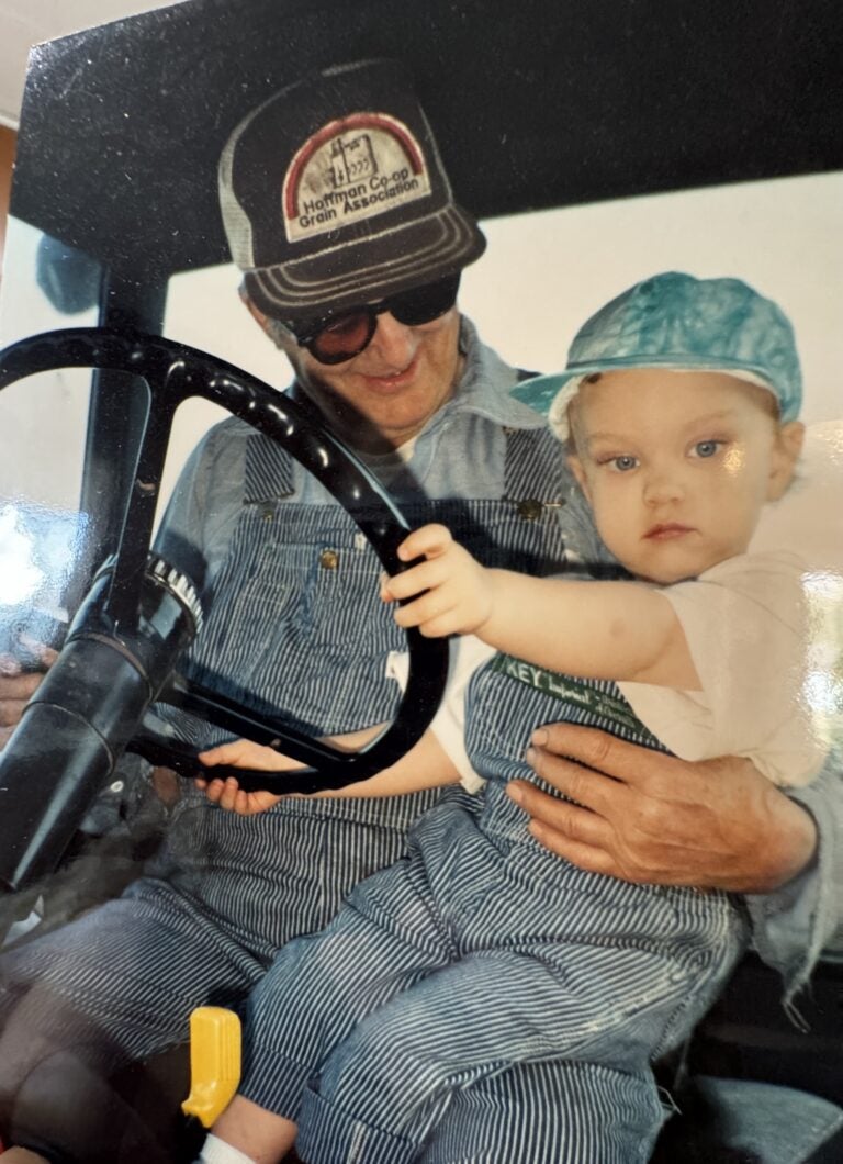 man in overalls and a cap holds a baby wearing a blue hat on his lap, sitting behind the steering wheel of a vehicle