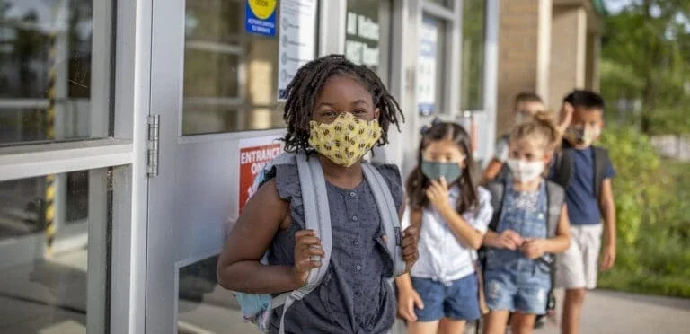 young boys and girls lined up outside of school wearing backpacks and masks.