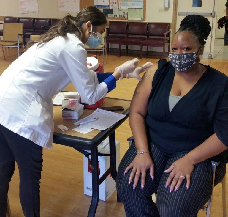 Image is of a woman sitting down getting a vaccine shot.