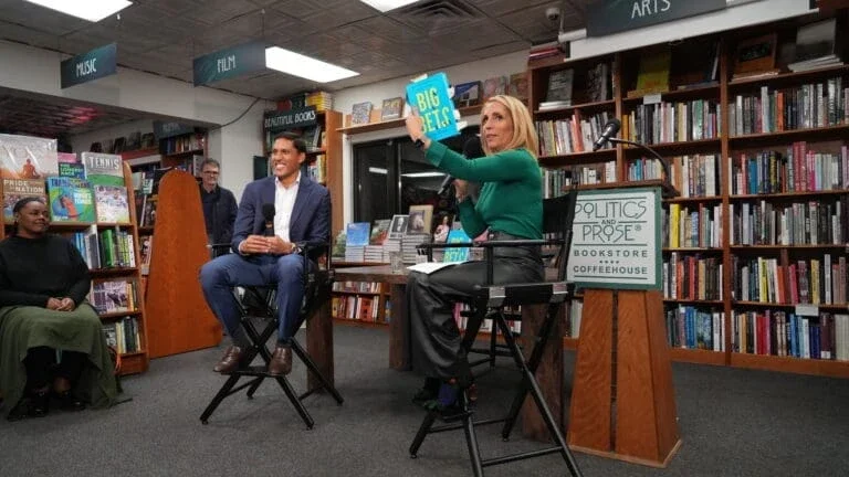 a man and woman sitting in chairs with the woman holding up a book towards a crowd