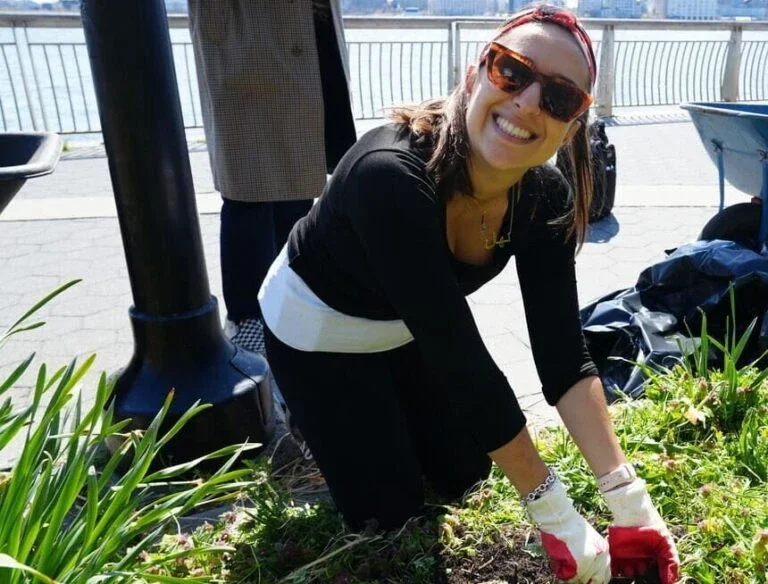 woman wearing a black shirt, white waistband, and red headband is smiling while gardening near a waterfront. She is kneeling, surrounded by greenery, with gloves on her hands, and sunglasses on her face