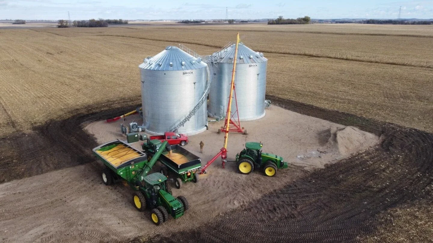 aerial view of grain silos on farmland with two tractors, one with a grain cart and the other with an auger. The surrounding land appears plowed