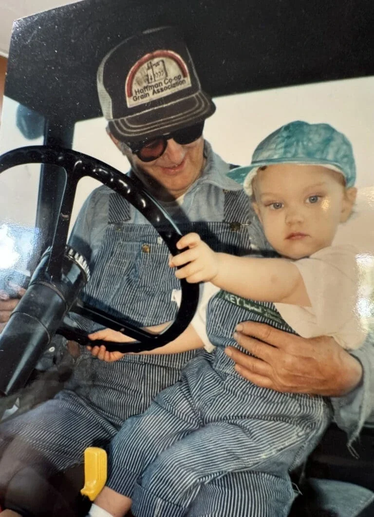 man in overalls and a cap holds a baby wearing a blue hat on his lap, sitting behind the steering wheel of a vehicle