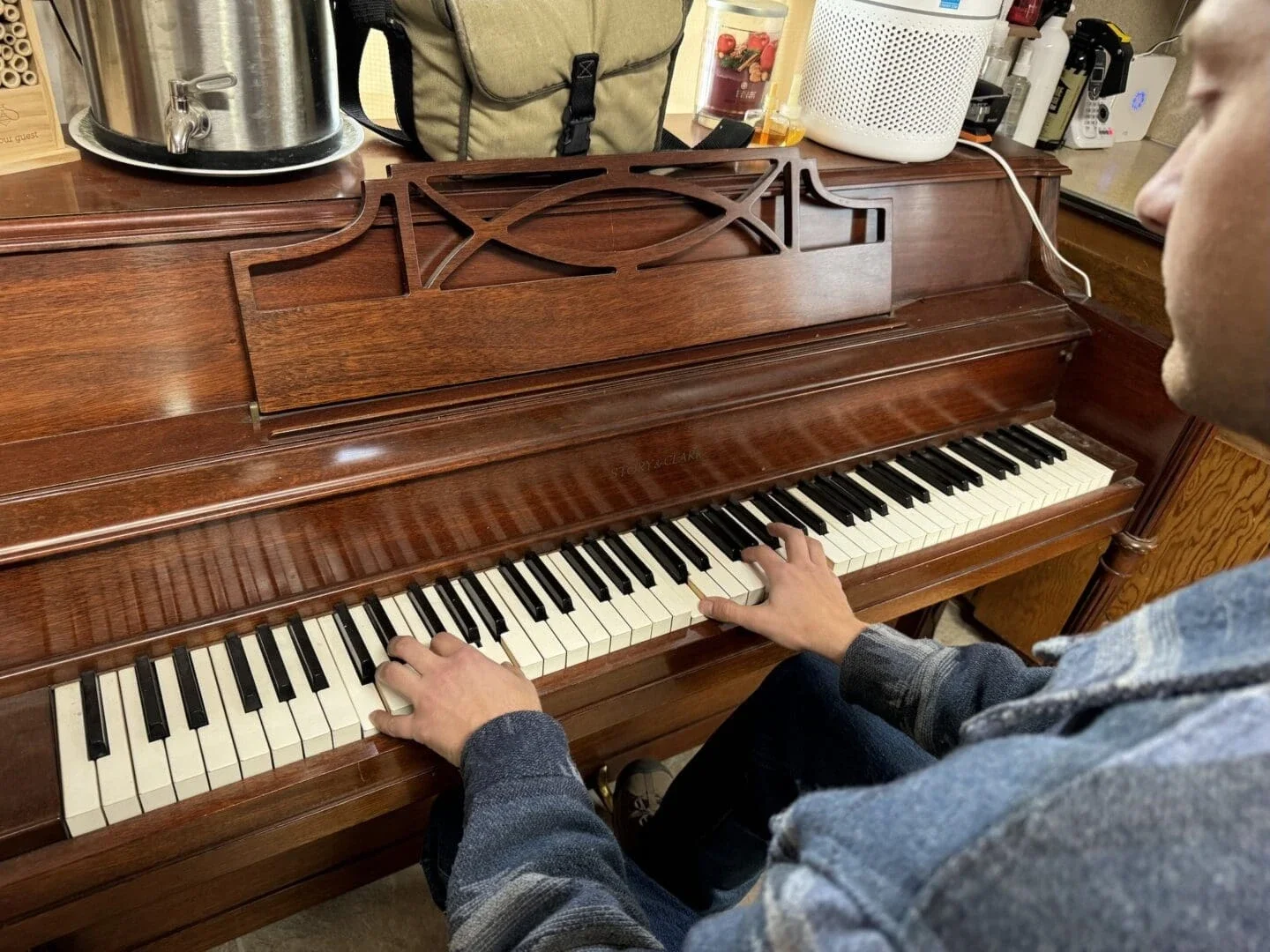 person playing a wooden piano with various items on top, including a speaker and a bag