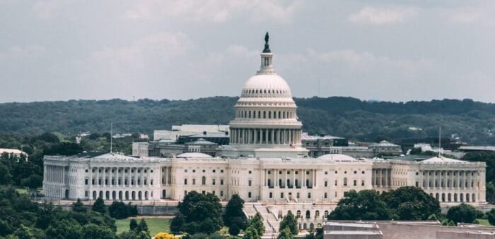 U.S. Capitol building under a cloudy sky, with surrounding greenery and distant hills